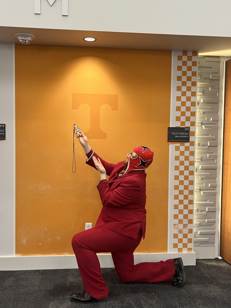 Man poses in front of a Power T, dressed in a monochrome red suit with matching luchador mask